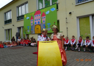 Taras przedszkolny, a na nim scenografia przedstawiająca zamek. Na tarasie siedzą przedszkolaki ubrane w stroje odpowiednie do przedstawienia. Na pierwszym planie dzieci przebrane za króla i królową.